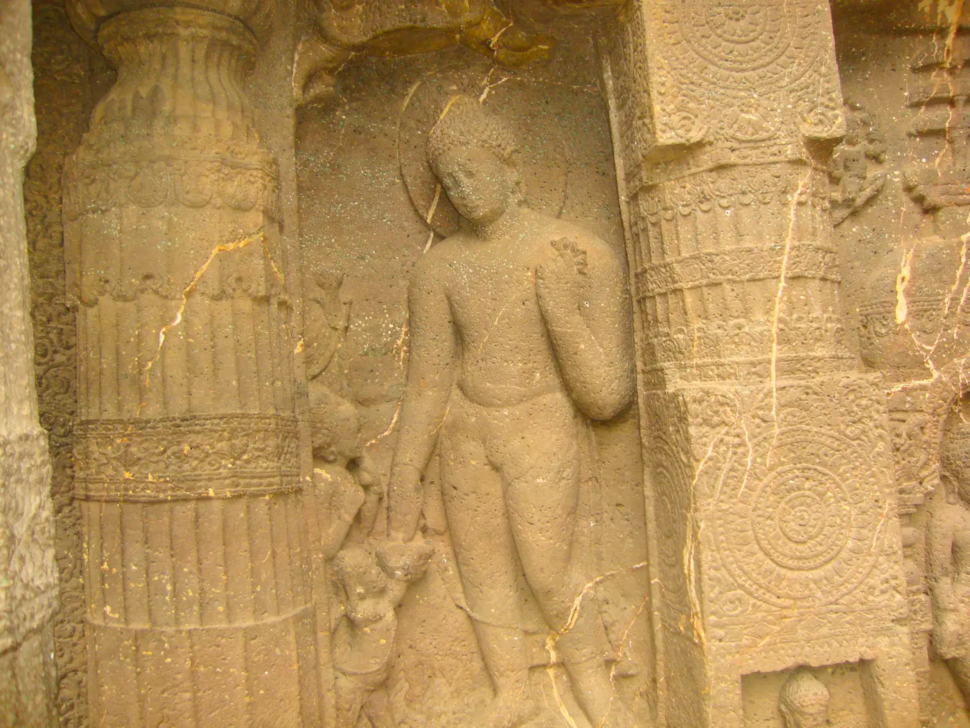 A weathered stone relief carved into a cave wall between two decorated pillars, showing a standing Buddha with a halo and a small child reaching up beside him