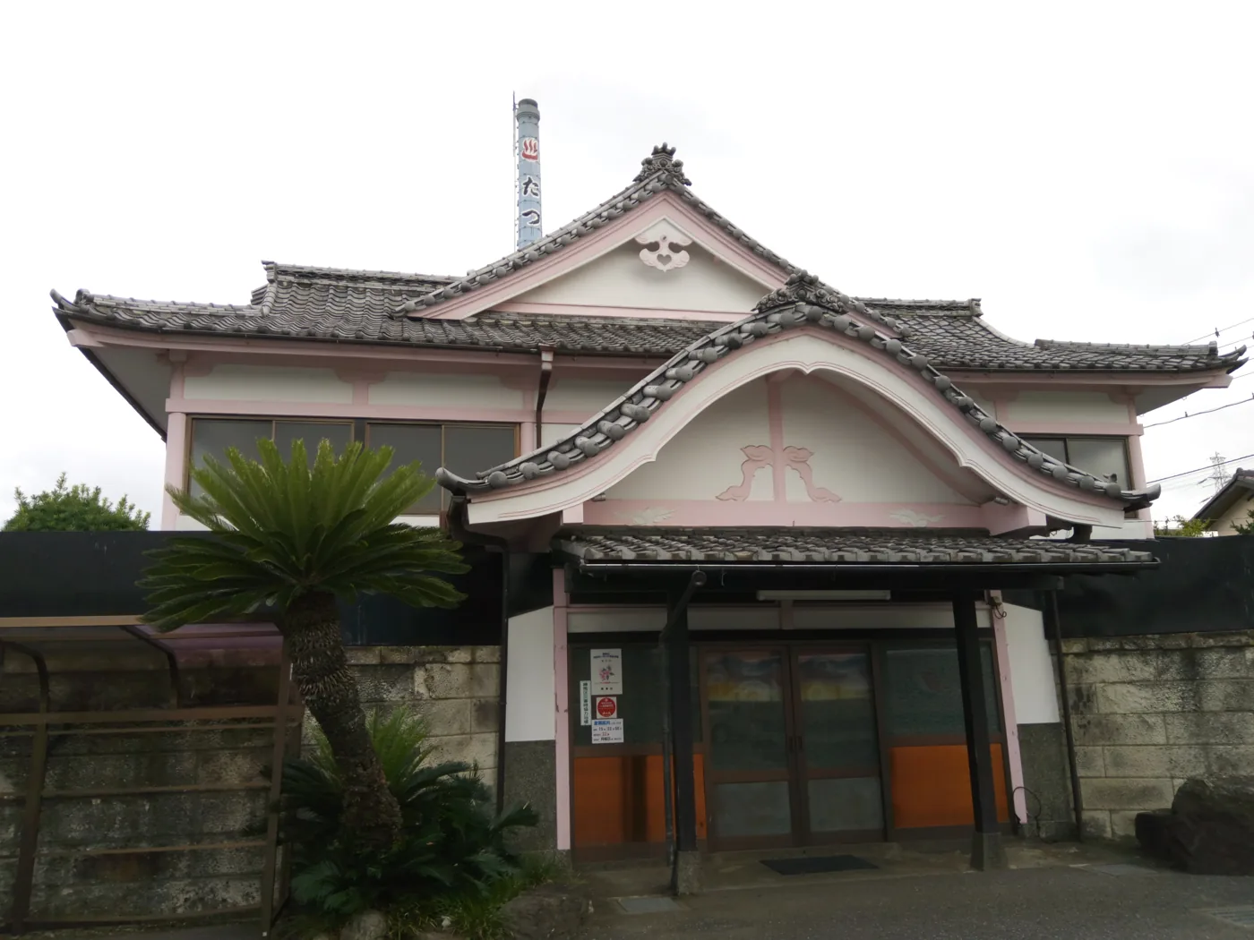 Tatsuno-yu, a Japanese neighbourhood public bath in Tokyo, built with a curved temple-style tiled roof, a tall chimney and a cycad palm by the entrance