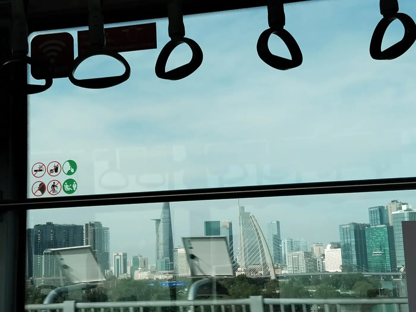 The Saigon River and the Ho Chi Minh City skyline seen through the window of a metro carriage, with hand straps above