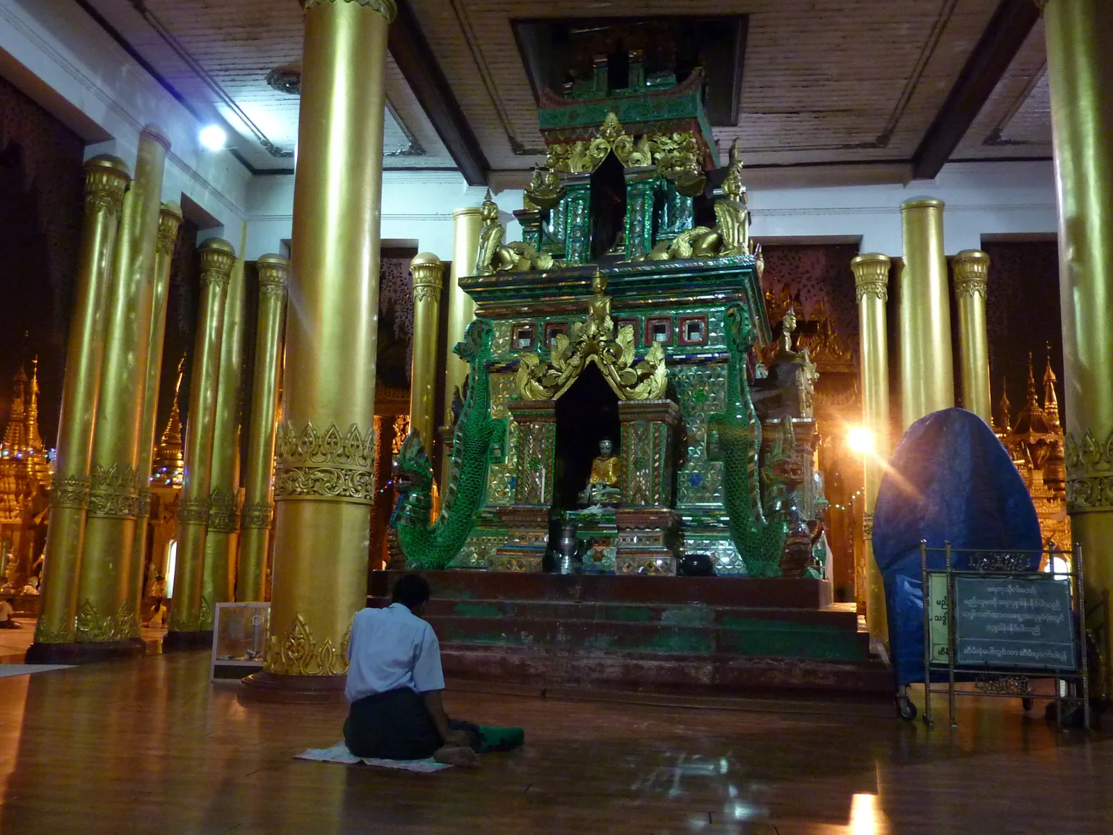 A person seen from behind, sitting alone on a mat on a polished floor before a gilded shrine with tall golden pillars at night