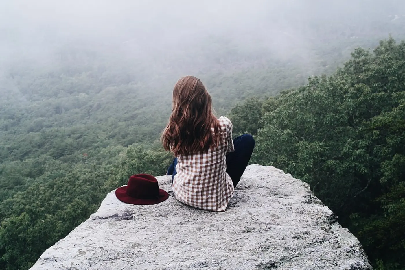A woman seen from behind sitting quietly on a rock above a misty green forest, a red hat beside her