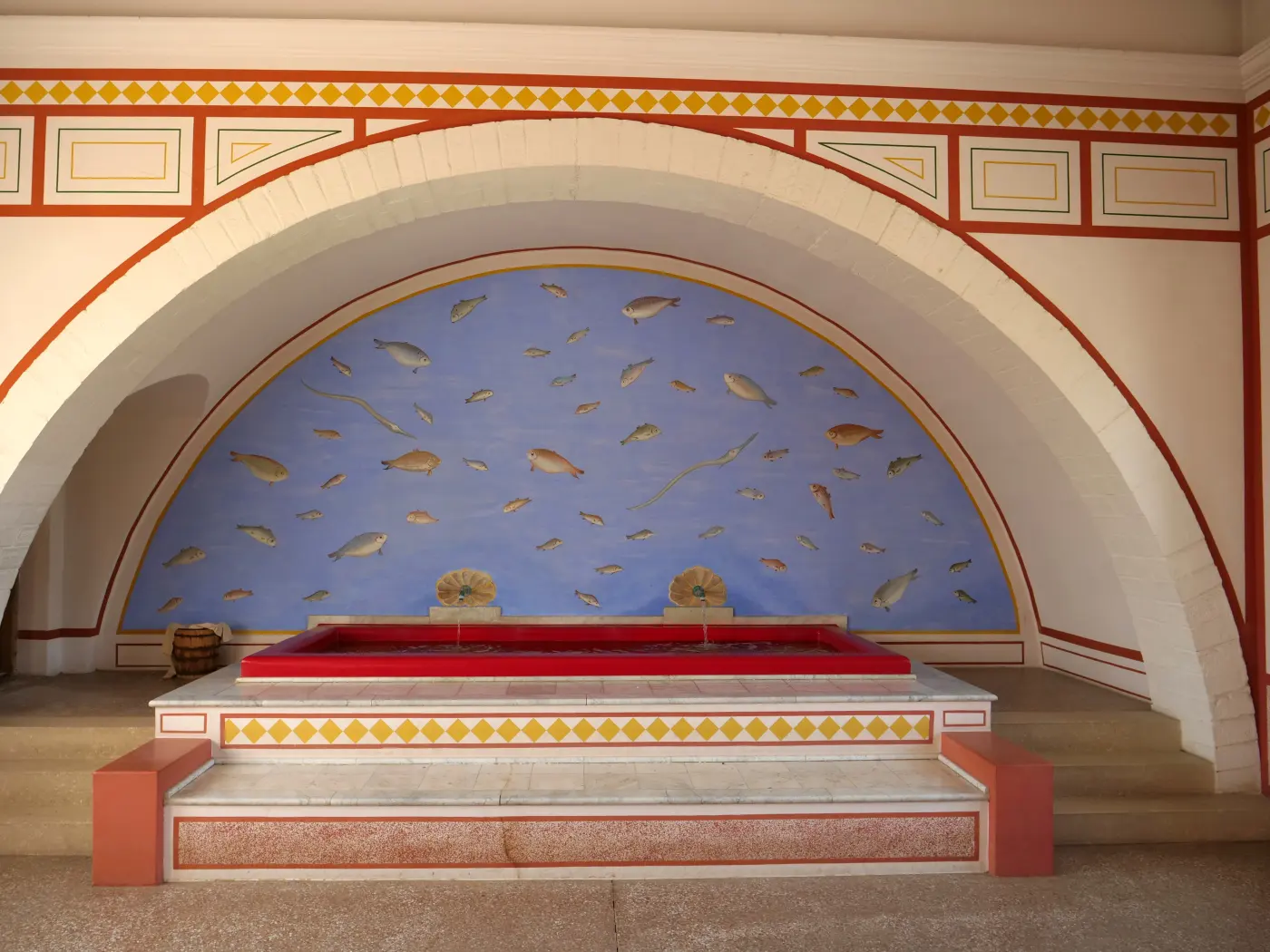Small cold-water pool under a painted arch with fish murals in a reconstructed Roman bathhouse