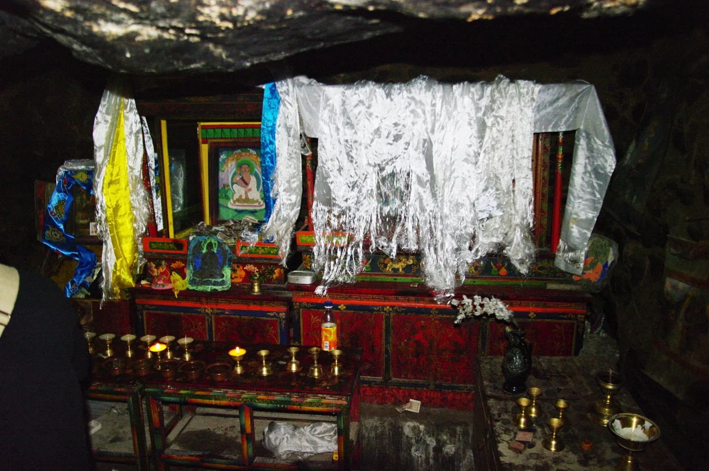 Inside Milarepa's meditation cave in Nyalam, Tibet: a low rock ceiling over a shrine with white offering scarves, butter lamps and a painted image of the yogi