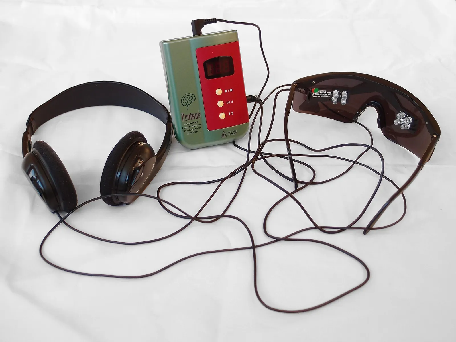 Small green control unit connected by cables to a pair of LED goggles and over-ear headphones on a white surface.