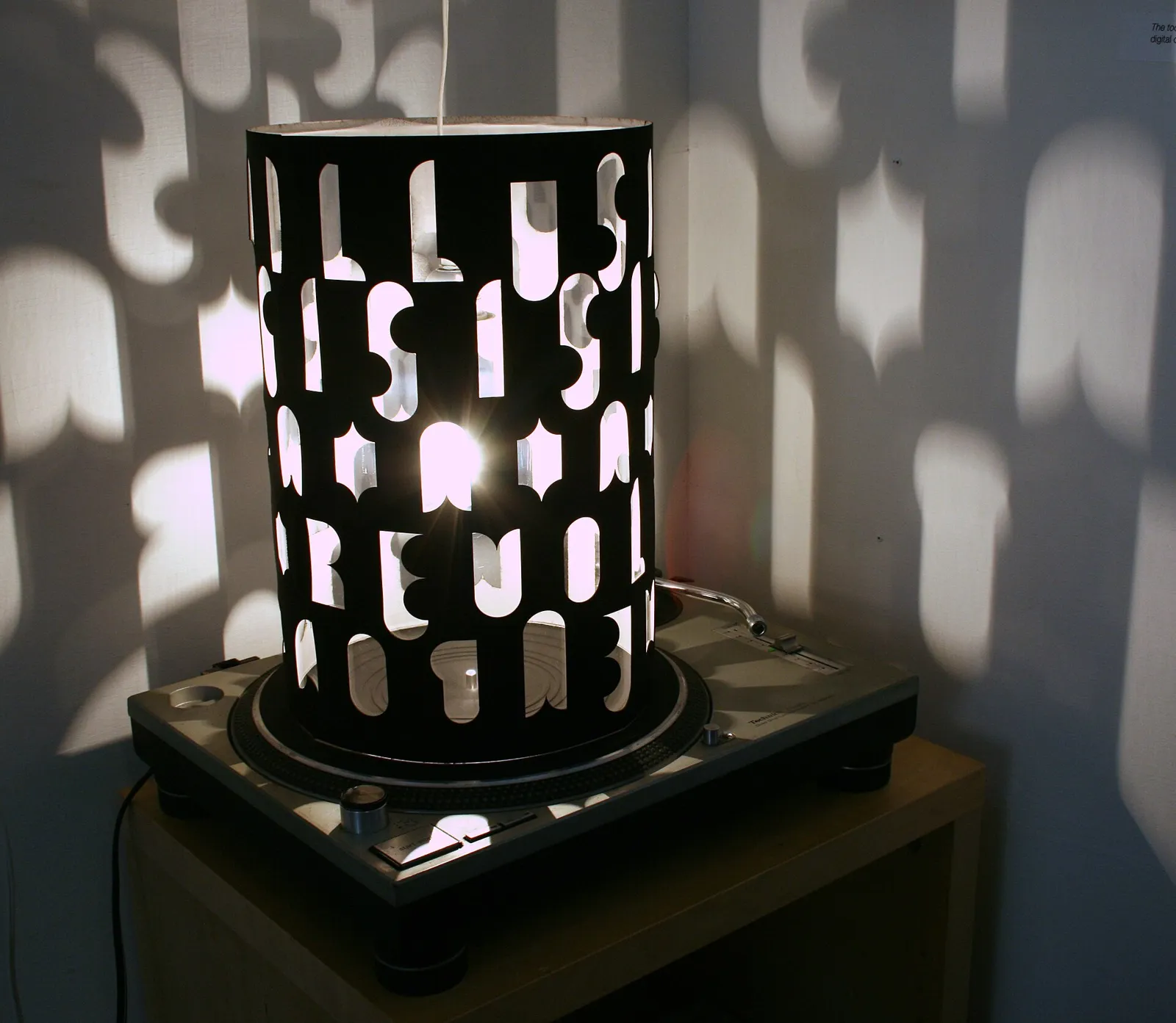 Black cylinder with cut-out patterns lit from inside, casting shapes of light onto the surrounding walls.