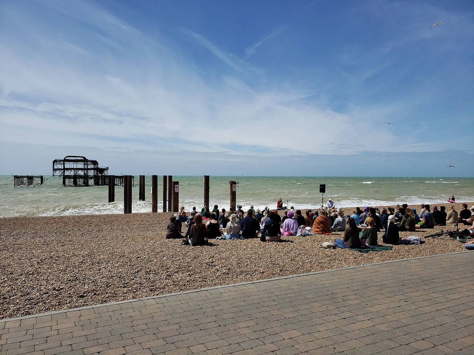 Group of people sitting quietly on a pebble beach facing the sea under a wide blue sky, an old pier in the distance.