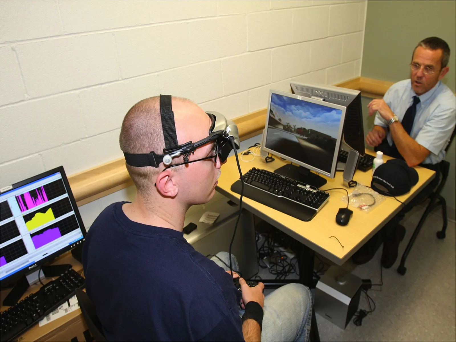 Man wearing a sensor headset in front of computer screens while a researcher watches