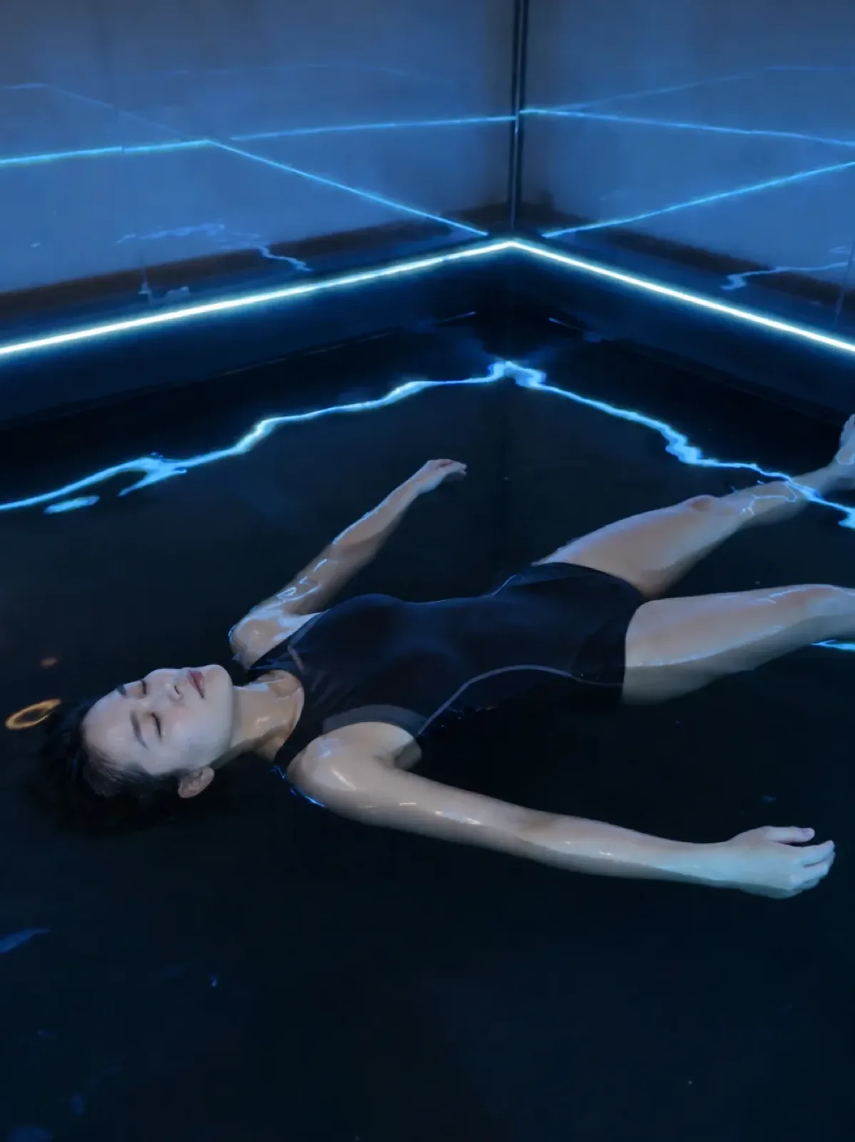 A woman in a black swimsuit floating on her back with her eyes closed and arms open, in dark water lit by soft blue light