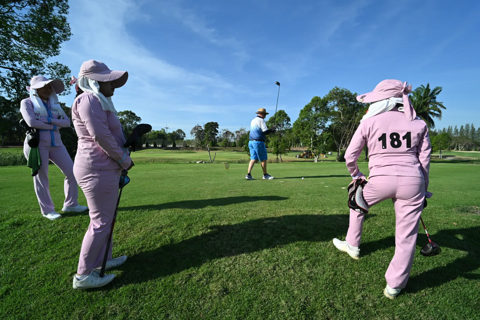 Caddies in pink uniforms and wide sun hats wait on the grass while a golfer in blue tees off under a clear sky