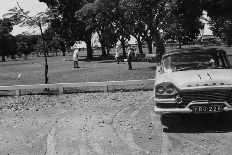 Black-and-white photo of golfers teeing off under tall trees at the Saigon Golf Club, with an old car parked in the foreground