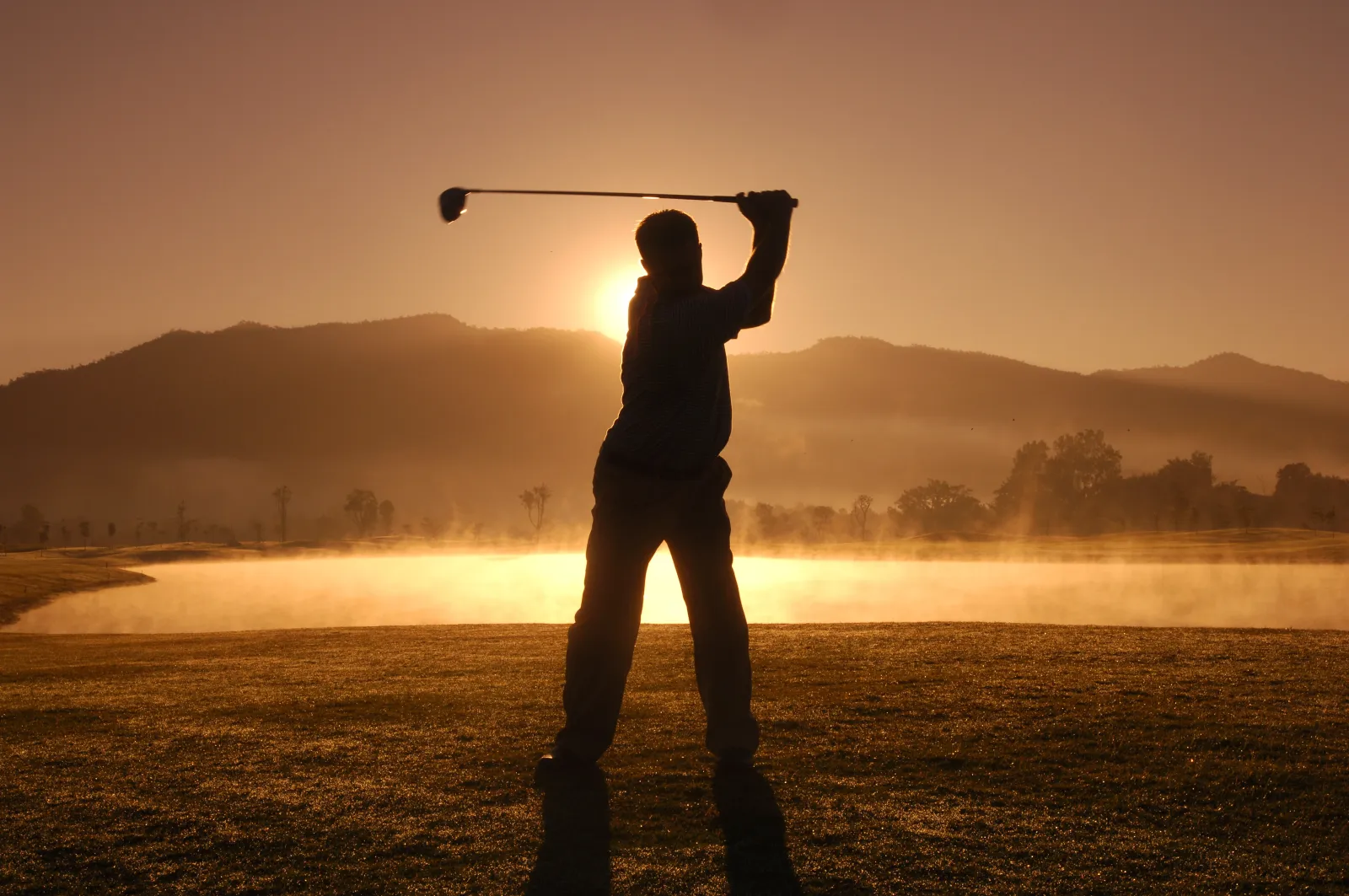 Silhouette of a golfer at the top of the backswing against a misty sunrise over a lake and hills