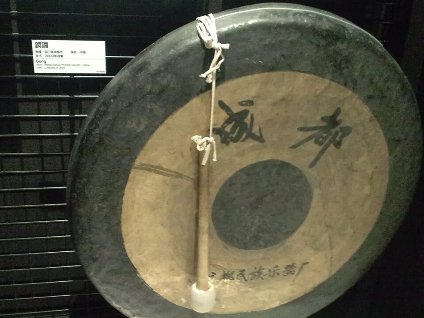 Un gong chino plano con el borde doblado hacia arriba y caracteres chinos pintados, colgado junto a su mazo en una vitrina de museo