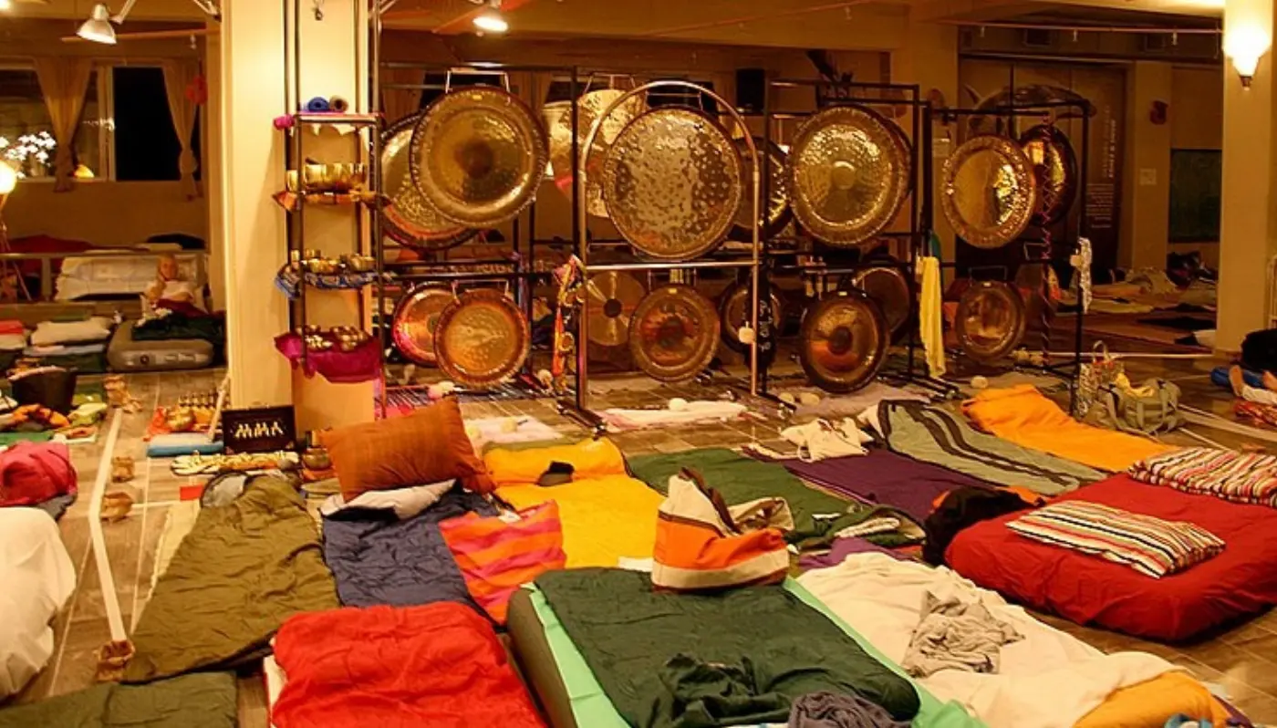Una sala preparada para un baño de gong, con una pared de gongs en sus soportes y esterillas y mantas de colores extendidas en el suelo