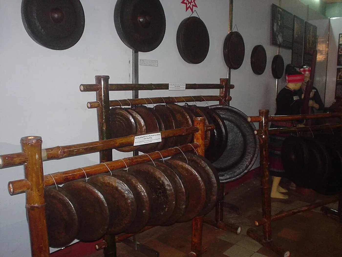 Hileras de gongs de bronce oscuro de las Tierras Altas Centrales sobre estantes de madera, con gongs más grandes colgados en la pared del fondo