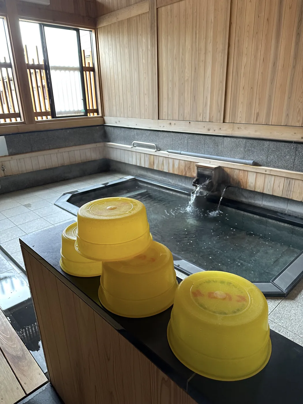 Nhà tắm onsen nhỏ bằng gỗ ở Nhật, nước suối nóng chảy vào bồn viền đá, phía trước xếp những chậu rửa màu vàng