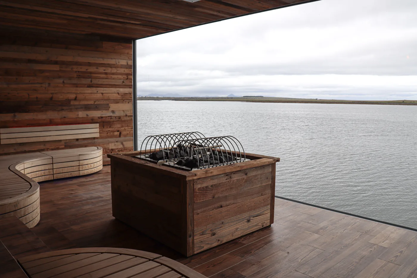 A Nordic-style sauna with curved wooden benches around a stove full of hot stones, and a wide window looking out over grey water