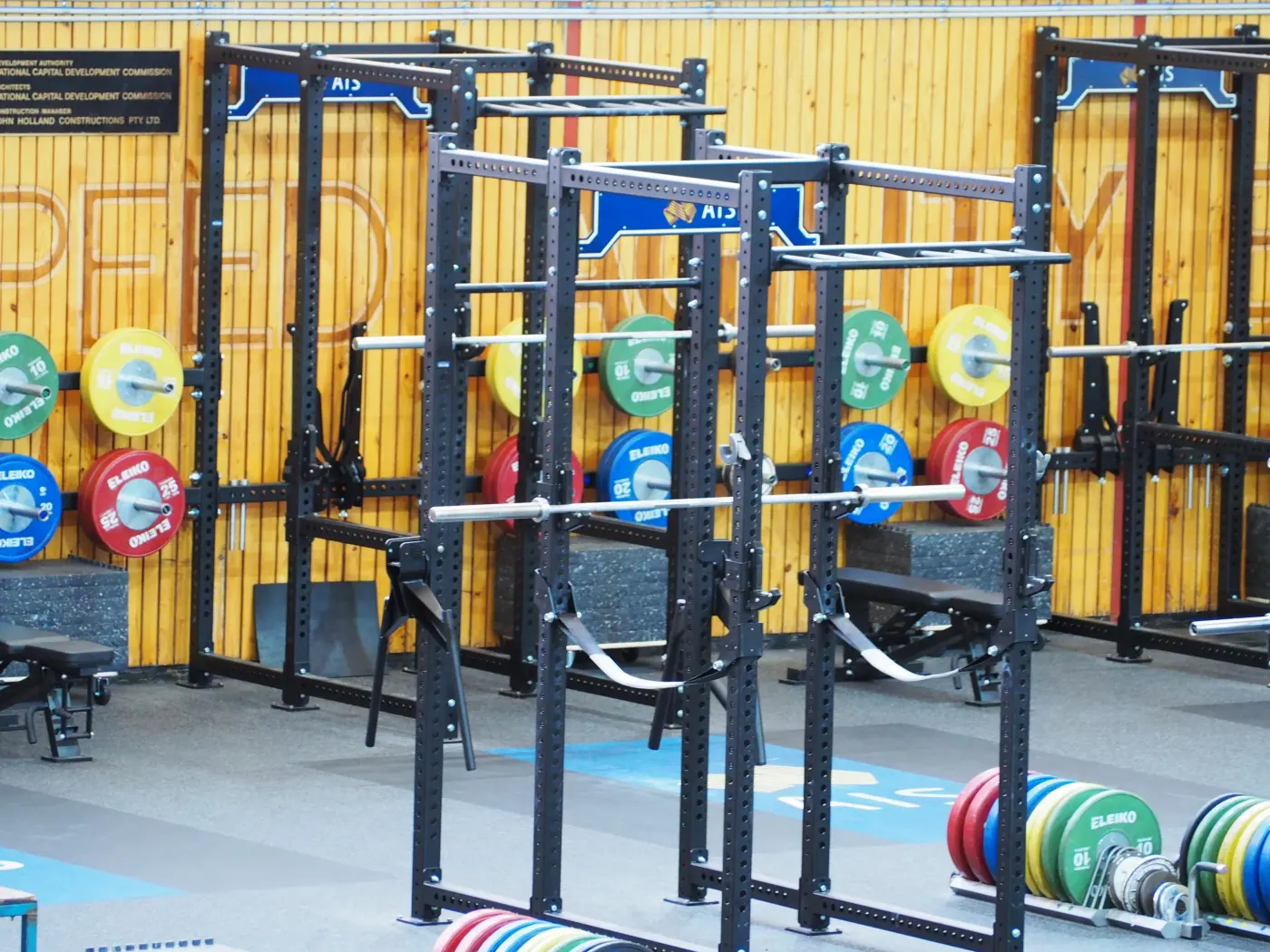 Rows of black squat racks, barbells and colourful weight plates in an empty strength gym