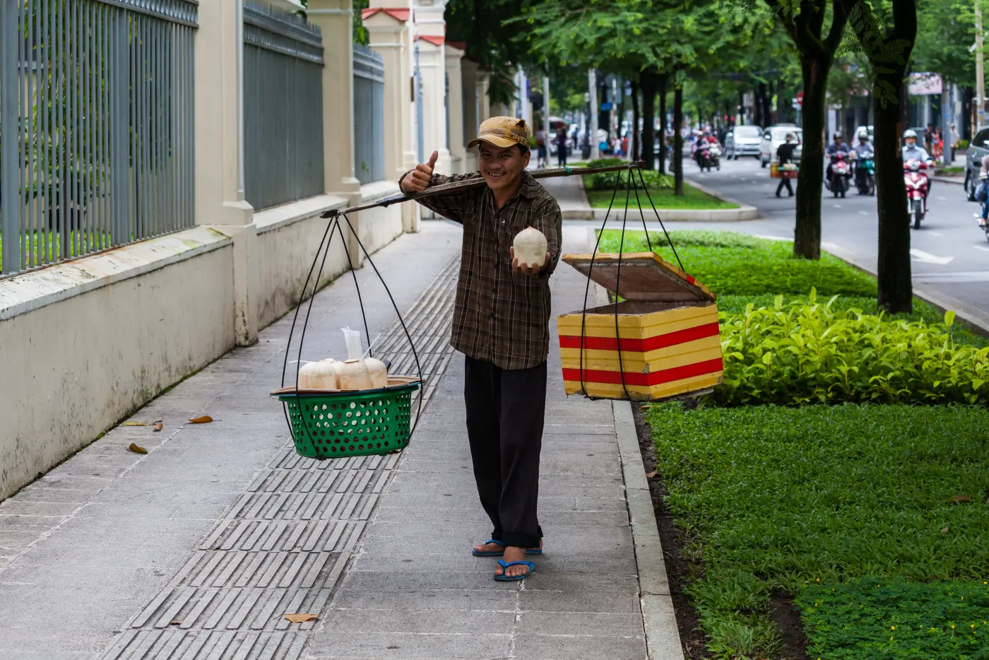 Coconut seller with a shoulder pole on a shady tree-lined pavement in Ho Chi Minh City