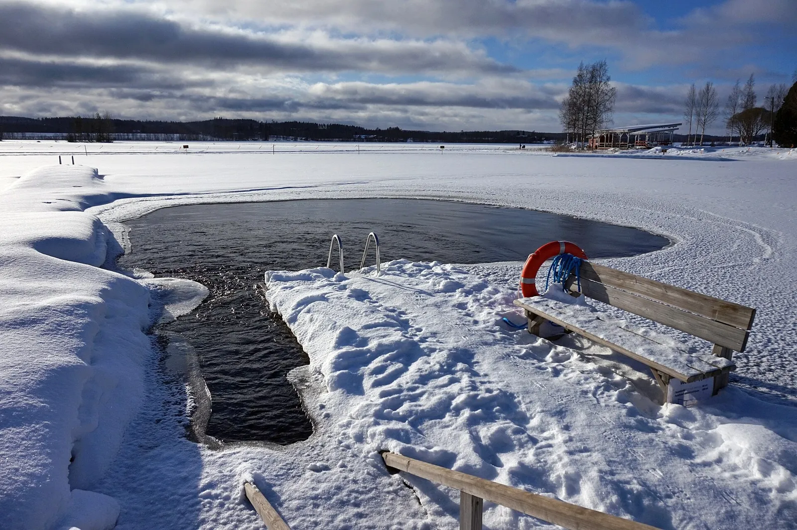 Rectangular hole cut in a frozen Finnish lake with a ladder and steps leading into dark water, snow-covered shore behind.