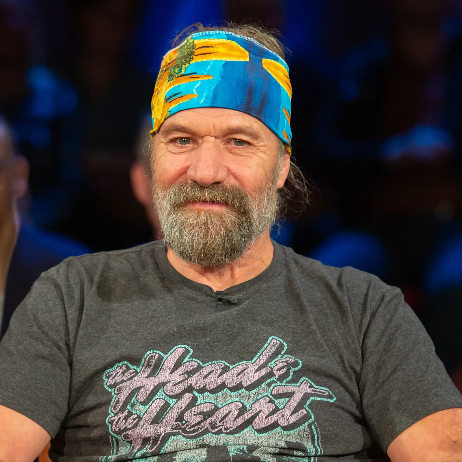 Portrait of Wim Hof, a bearded man with a colourful headband and grey T-shirt, smiling on a TV talk-show set.