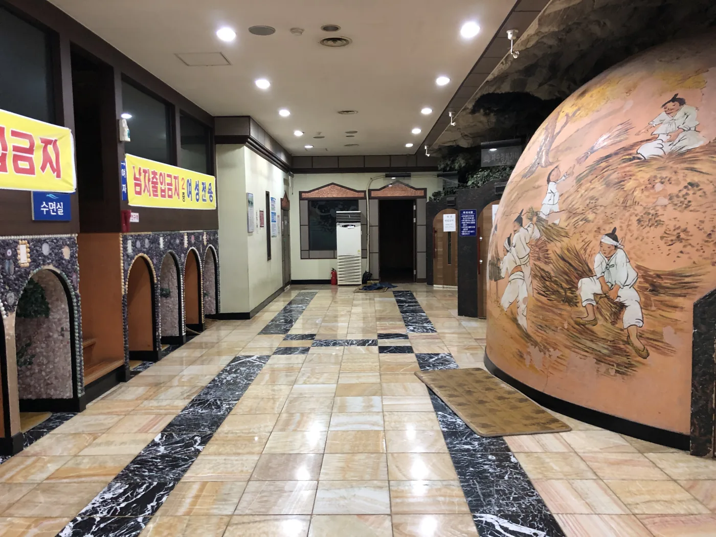 The corridor of a Korean jjimjilbang with a domed bulgama kiln room painted with a traditional mural, and a mat in front of its small door