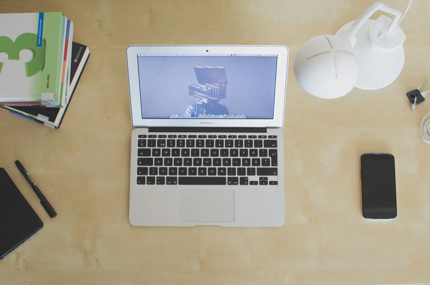A laptop, phone, notebooks and a desk lamp on a plain wooden desk, seen from above