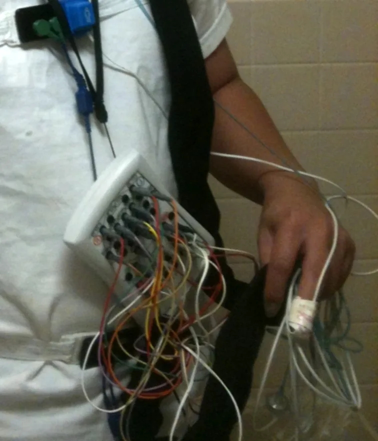 A person in a white shirt wired up with many coloured sensor leads running to a small recording box for an overnight sleep study