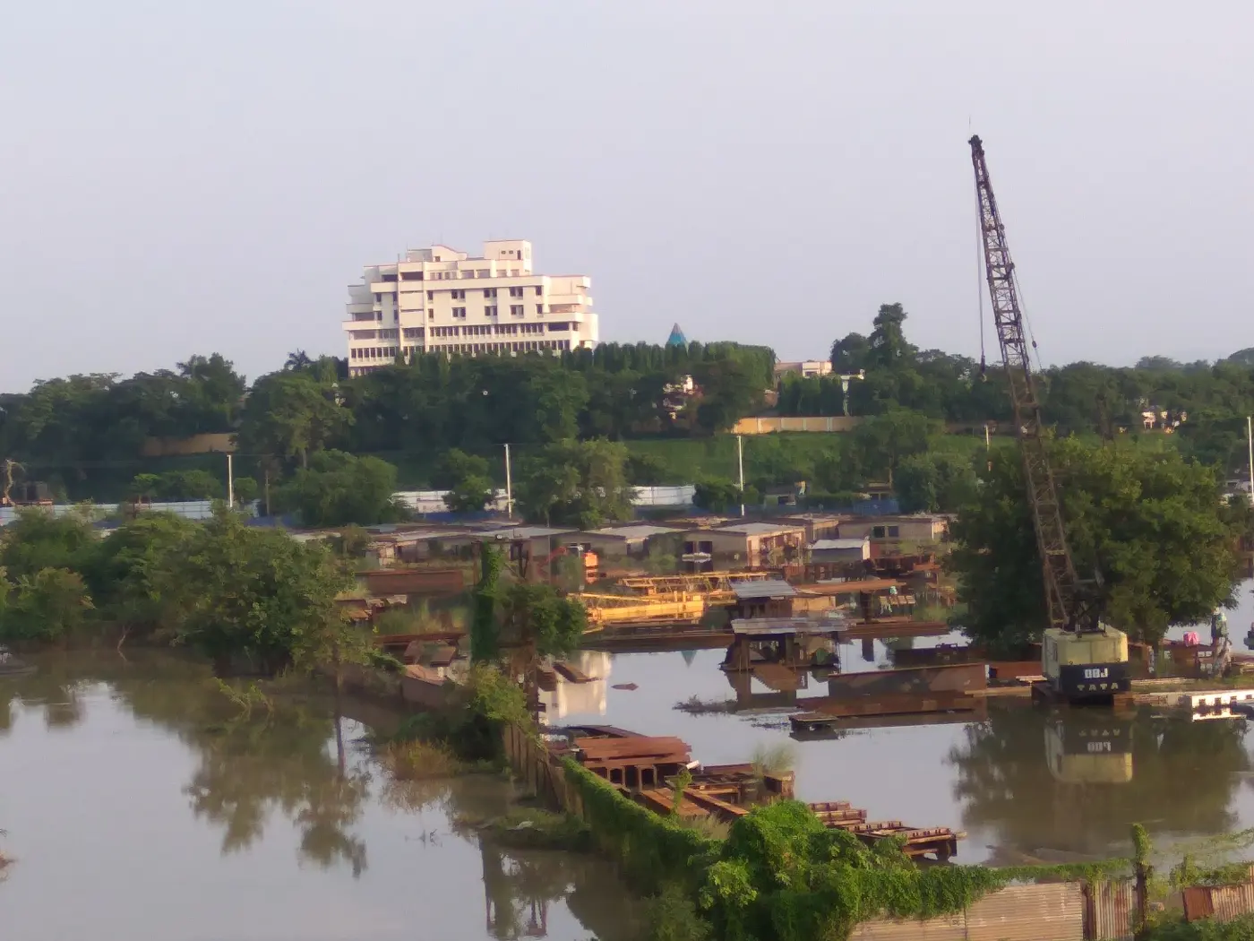 A white building on a green hill above a wide river, with boats and a crane in the foreground