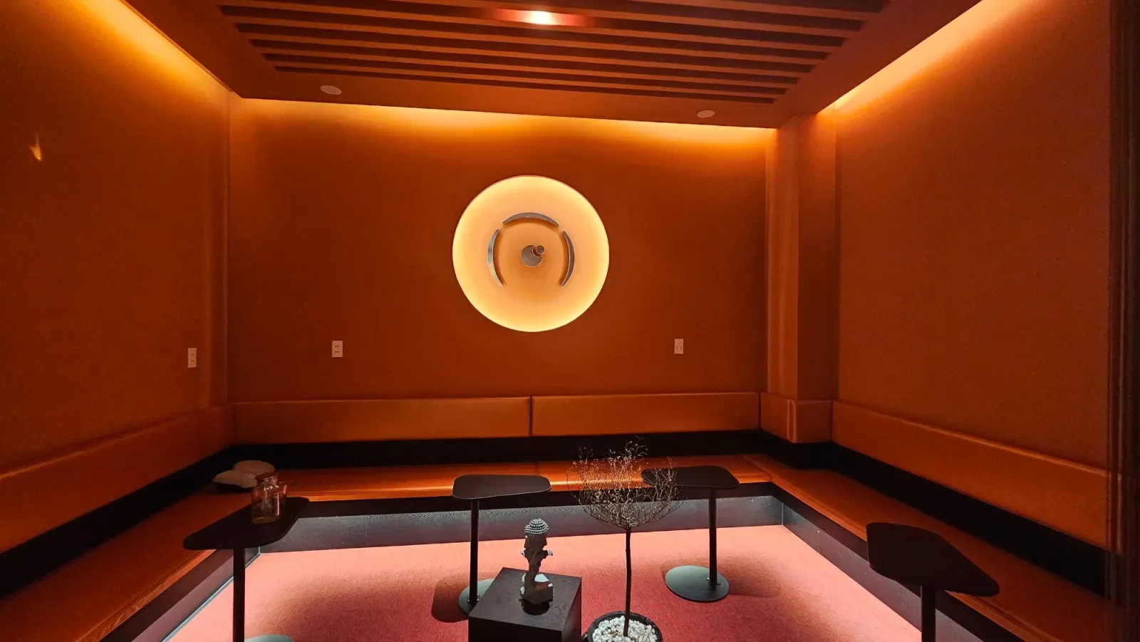 An empty orange lounge at Ōmni Space with low benches along the walls, small round tables, a stone head and a bare little tree, under a glowing round logo