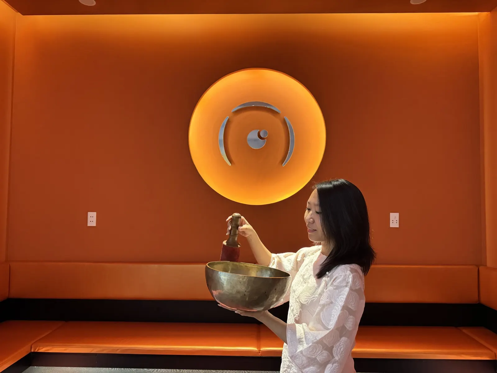 Una practicante sostiene un gran cuenco tibetano de latón y un mazo en un cálido salón naranja con una luz redonda que brilla en la pared