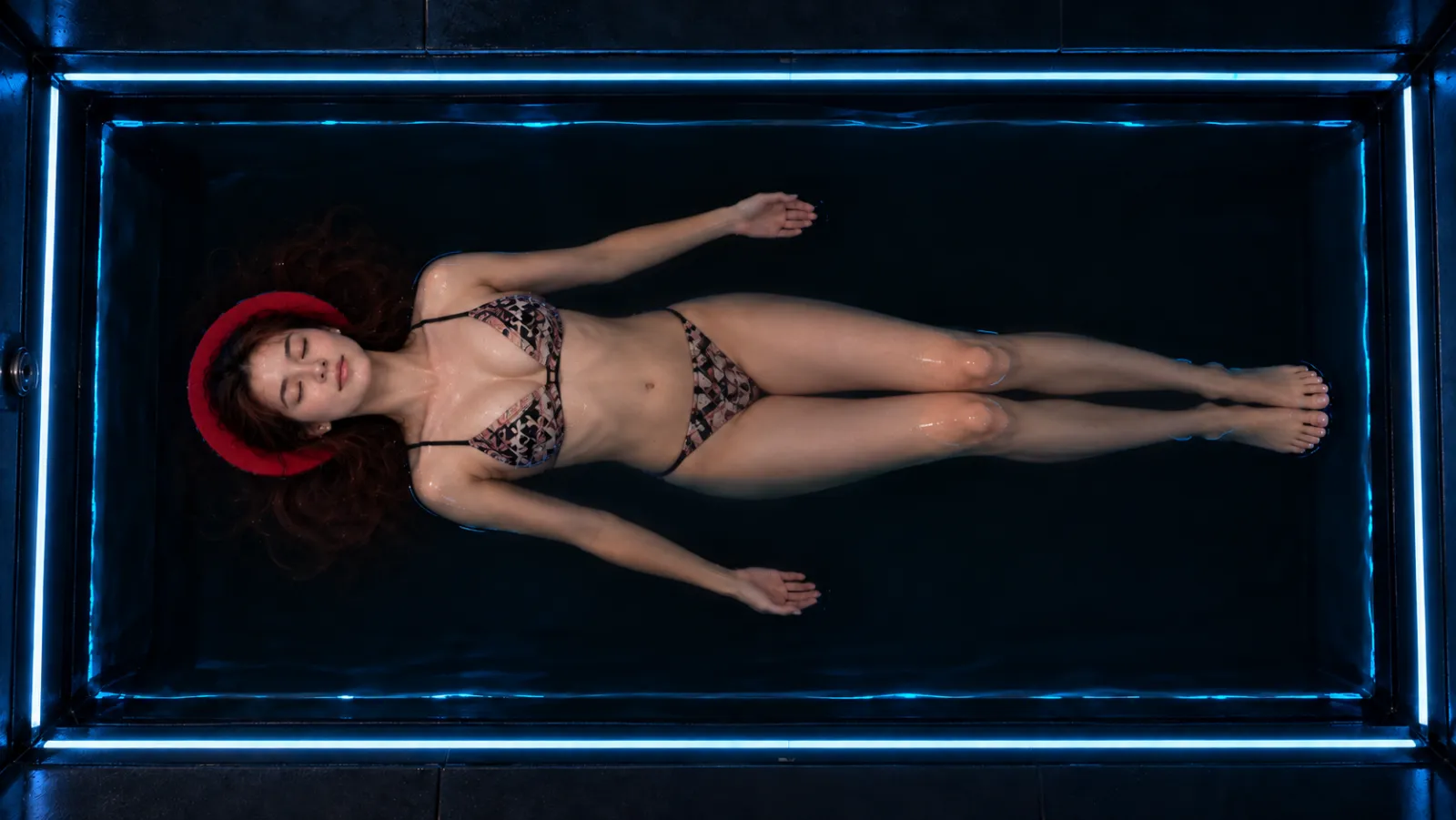 Seen from above, a woman in a patterned two-piece floating on her back in a rectangular float pool, arms loose at her sides, head on a red neck pillow, blue light along the edges