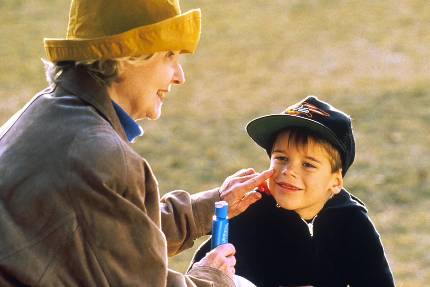 An older woman in a yellow sun hat smiling as she dabs sunscreen onto a young boy's cheek outdoors