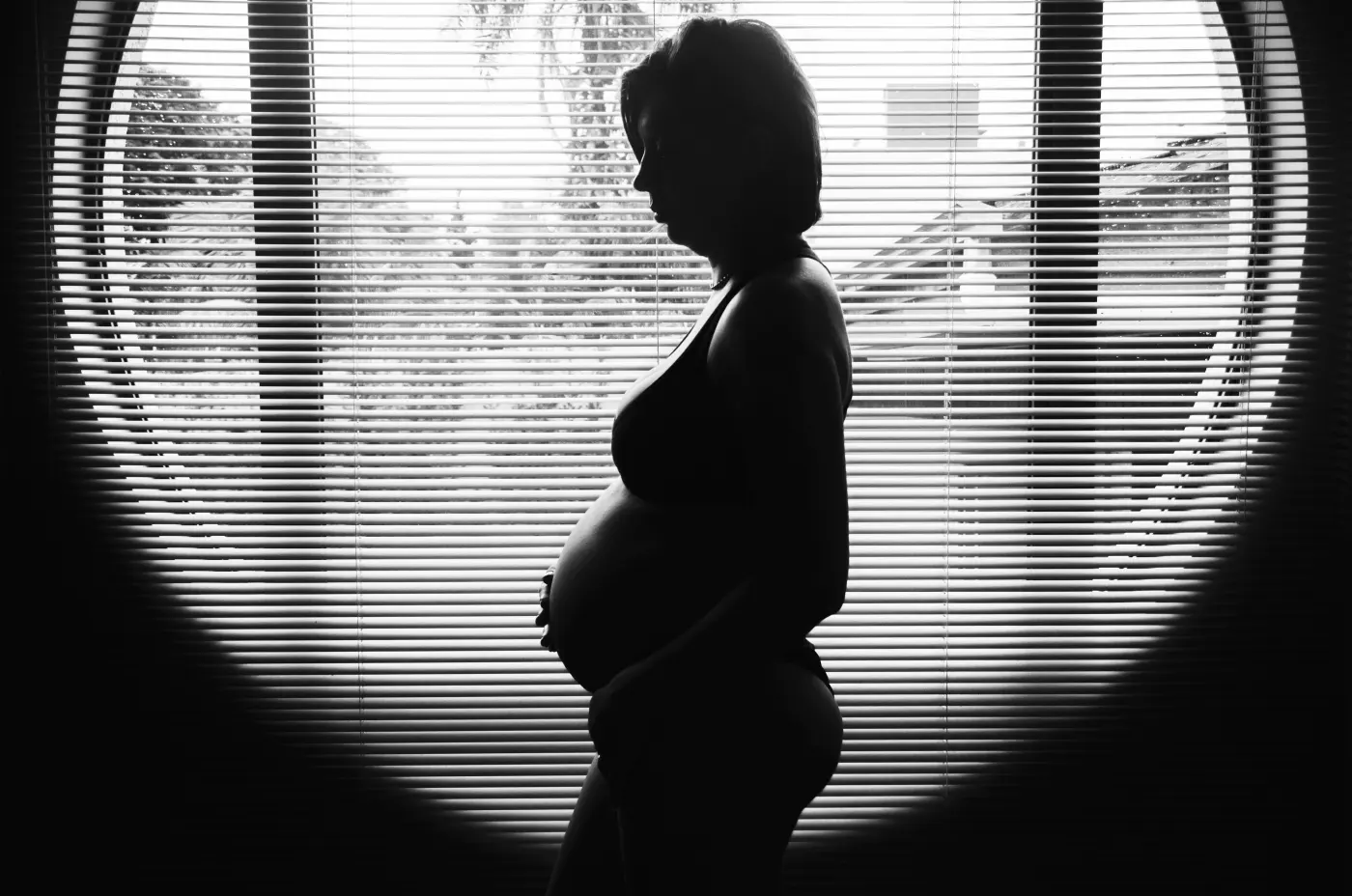 Black-and-white silhouette of a pregnant woman in profile standing by a round window with blinds