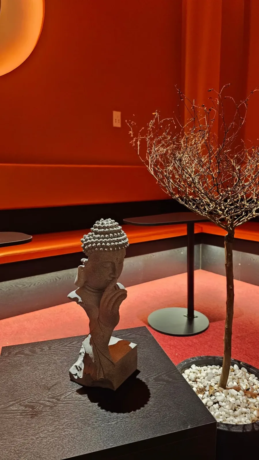 A small stone Buddha head resting its chin on one hand, on a dark table beside a bare decorative tree, in the orange lounge at Ōmni Space