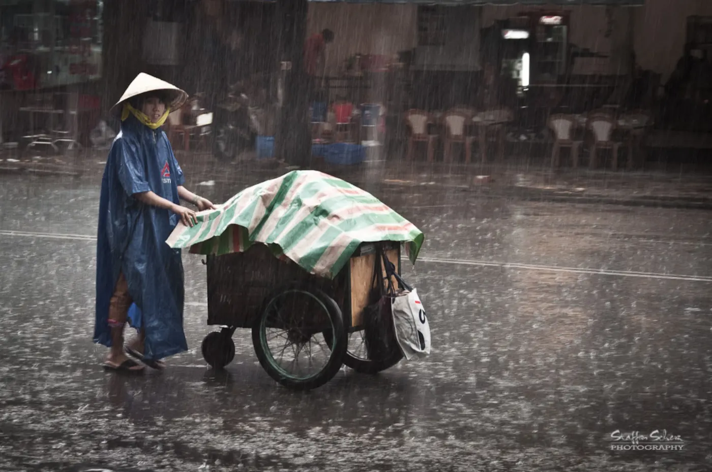 A street vendor in a blue raincoat and conical hat pushing a covered cart through heavy rain in Saigon