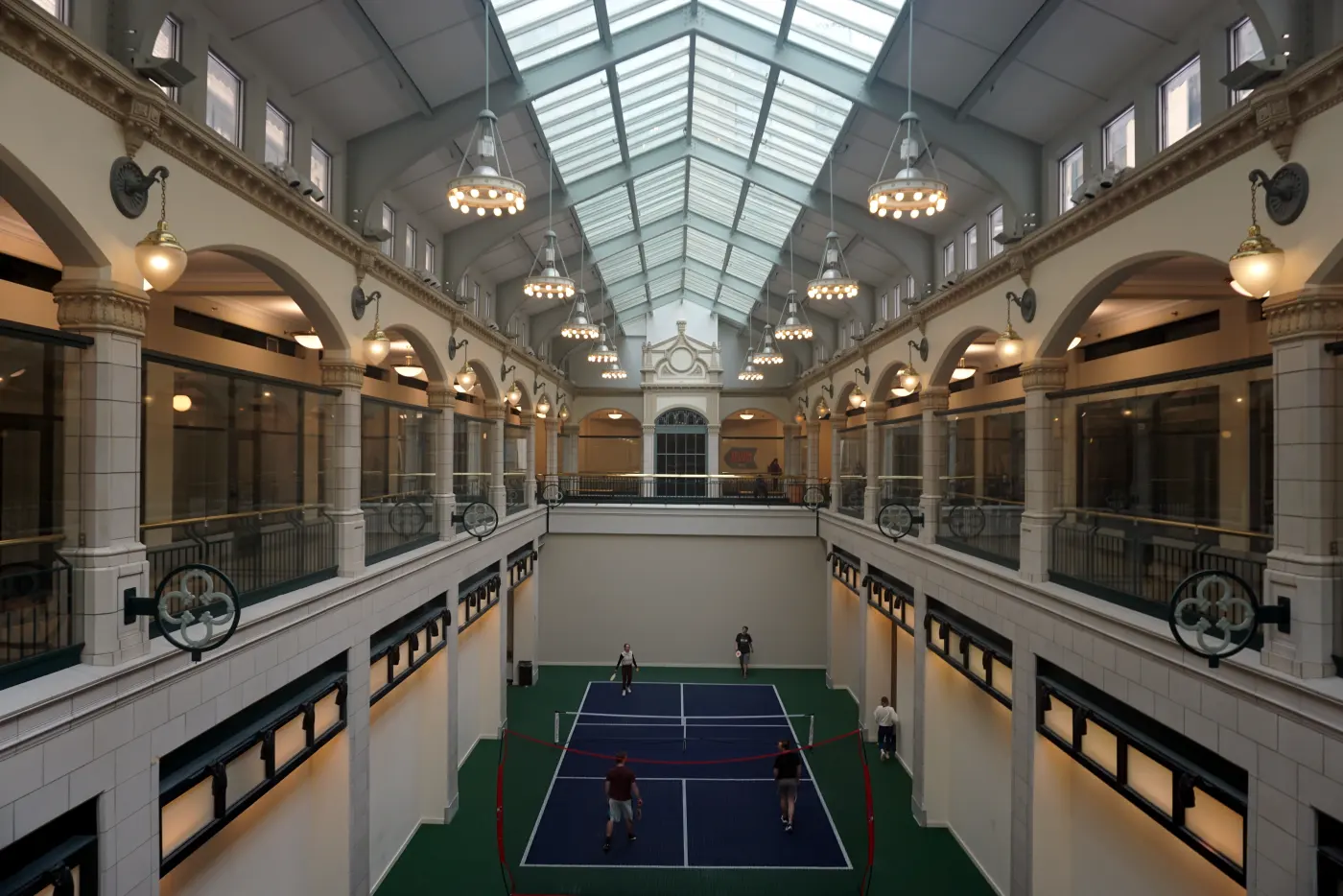 Players on an indoor pickleball court under a glass roof, seen from a balcony