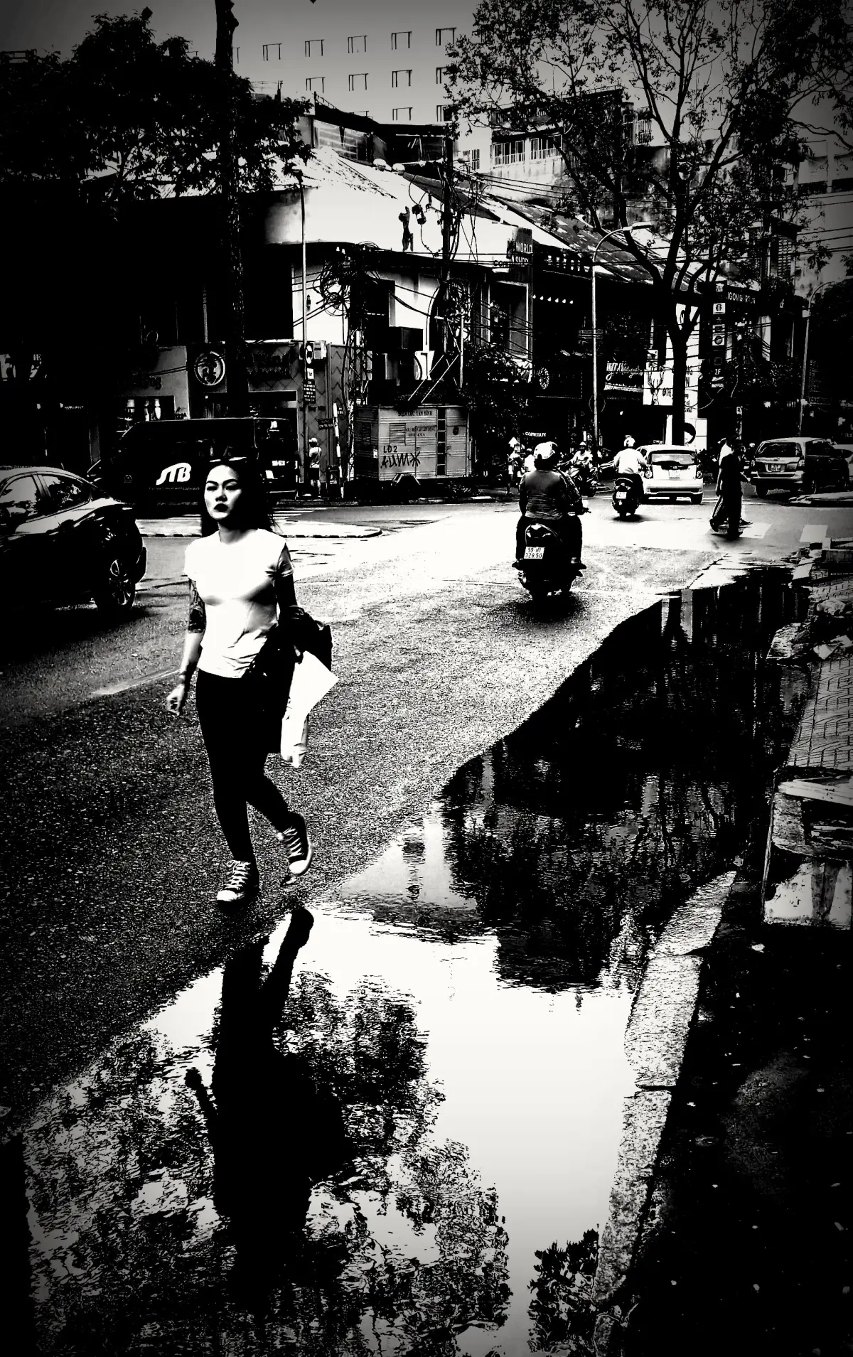 Black-and-white street in Ho Chi Minh City after rain, puddles reflecting trees and motorbikes