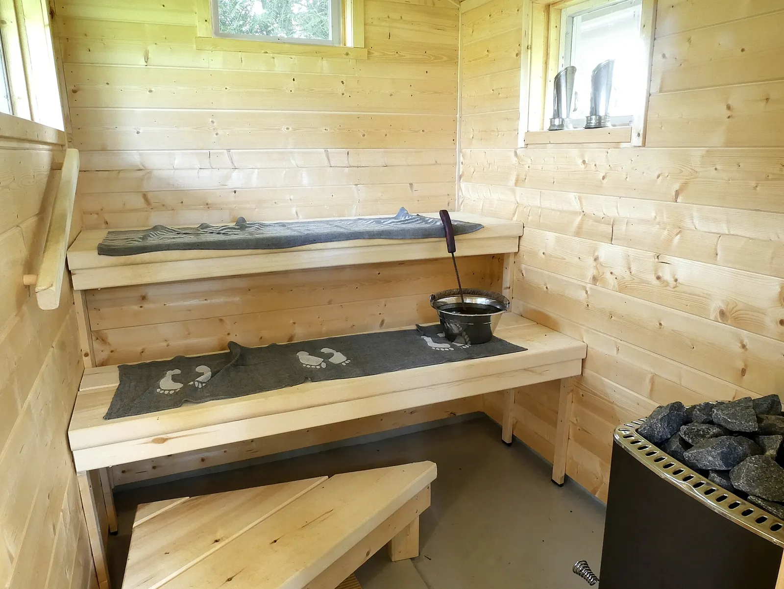 Light pine-panelled Finnish sauna room with two-level wooden benches and a stone-topped heater in the corner.
