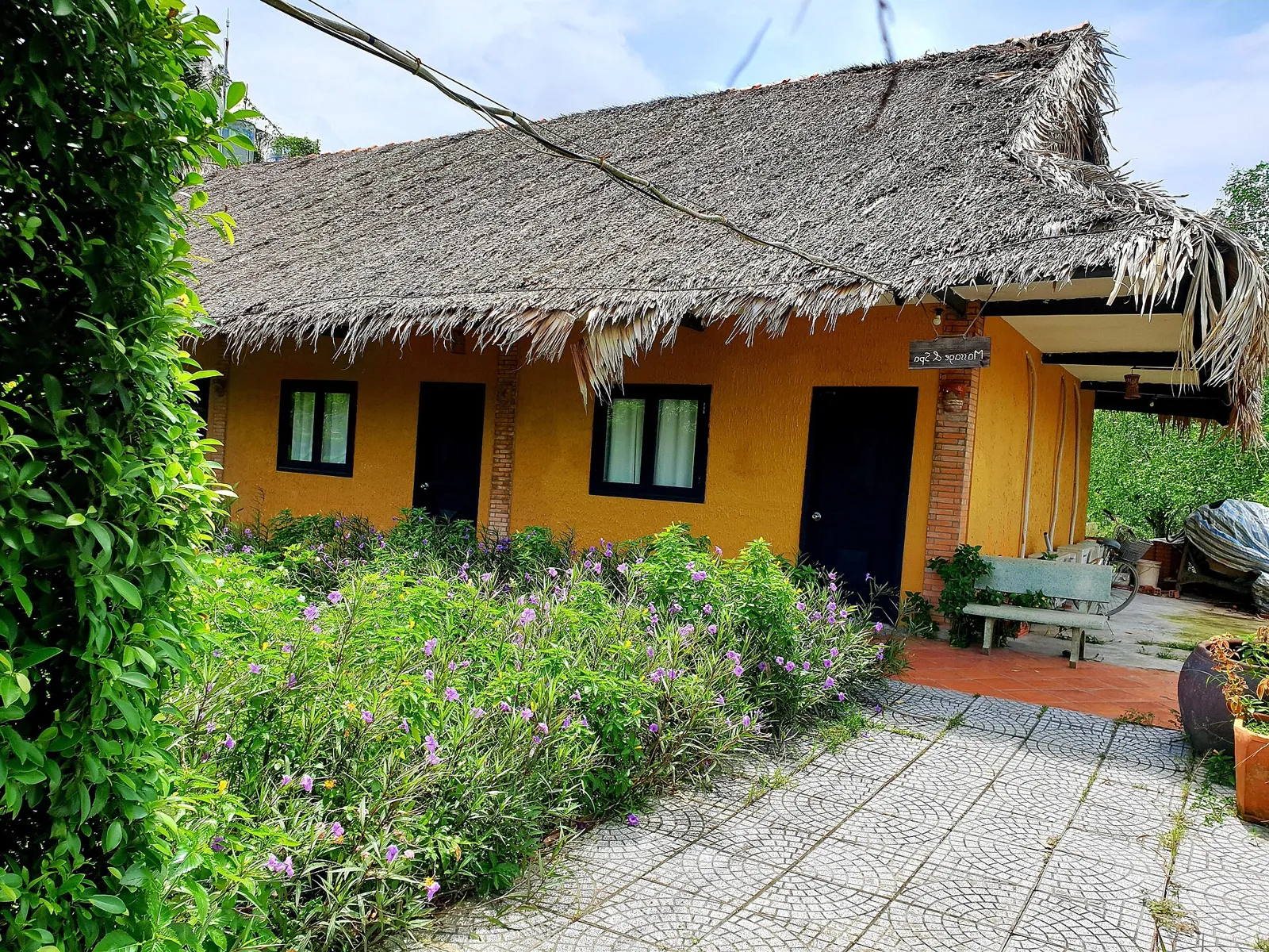 Small yellow house with a thatched palm-leaf roof surrounded by green plants and a paved garden path.