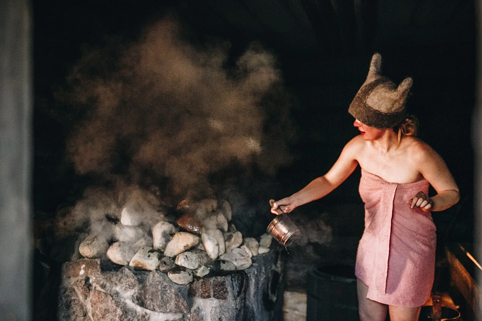 Woman in a towel and felt hat pouring water onto hot sauna stones, steam rising