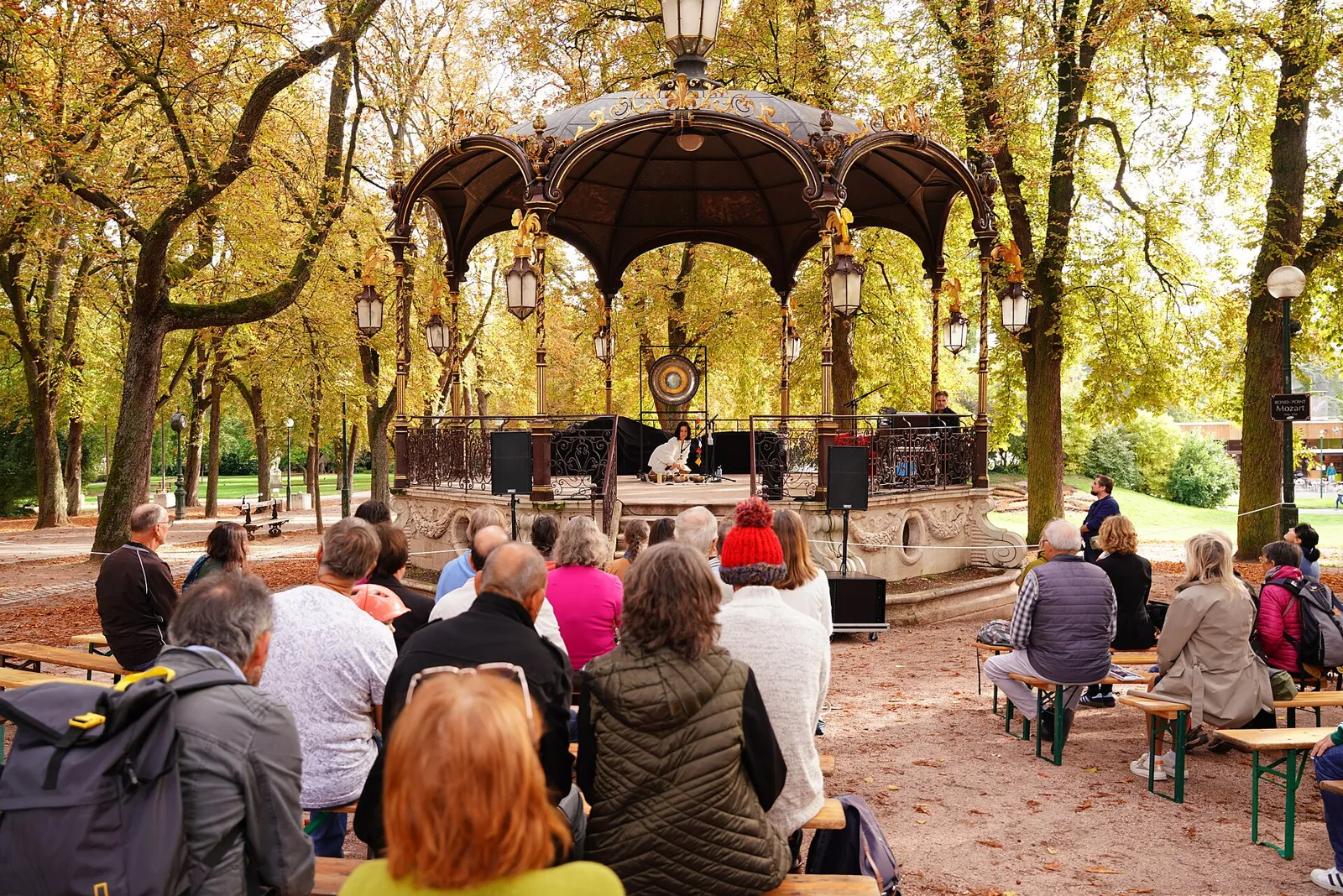 Audience seated on benches under autumn trees, listening to gongs played in an ornate park bandstand.