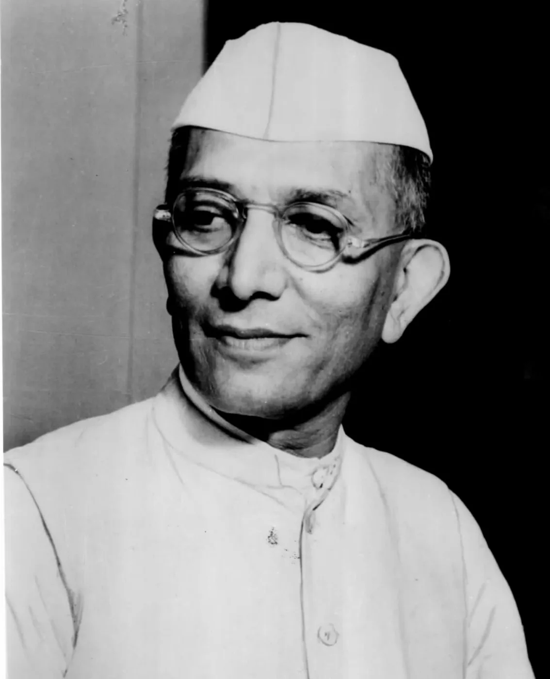 Black-and-white portrait of Morarji Desai, an elderly man in round glasses and a white Gandhi cap, smiling slightly