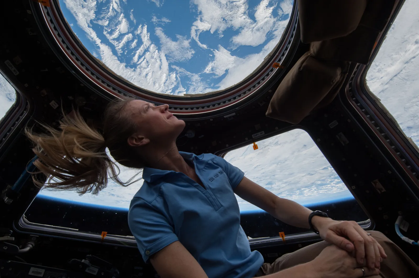 An astronaut in a blue shirt looks up at the Earth through the round windows of the space station, her hair floating freely around her head