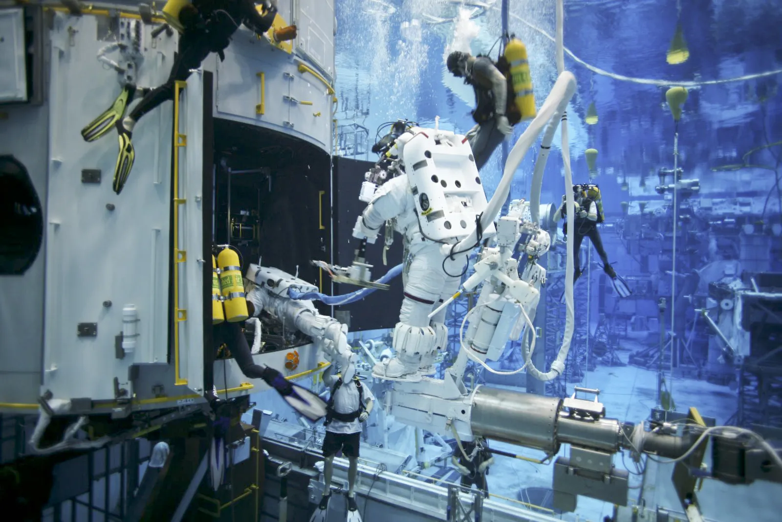 An astronaut in a white spacesuit works on a large model of the Hubble telescope underwater, with safety divers around him in a huge blue pool