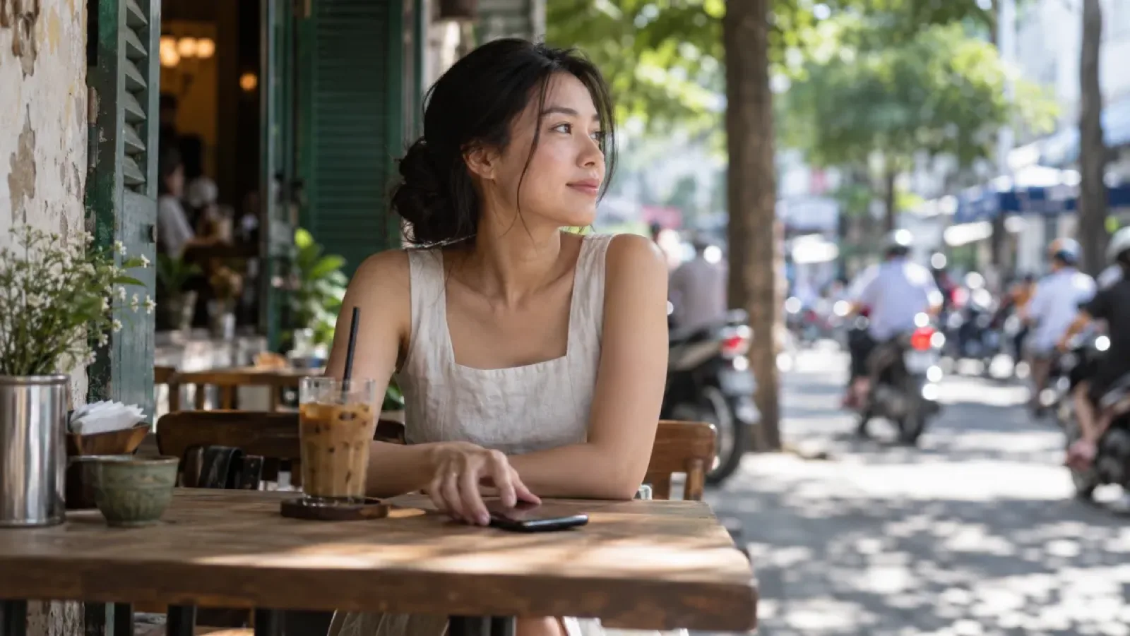 A young woman at a wooden table outside a Saigon sidewalk café, a glass of iced coffee in front of her, her phone face down under her hand, motorbikes passing in the street behind