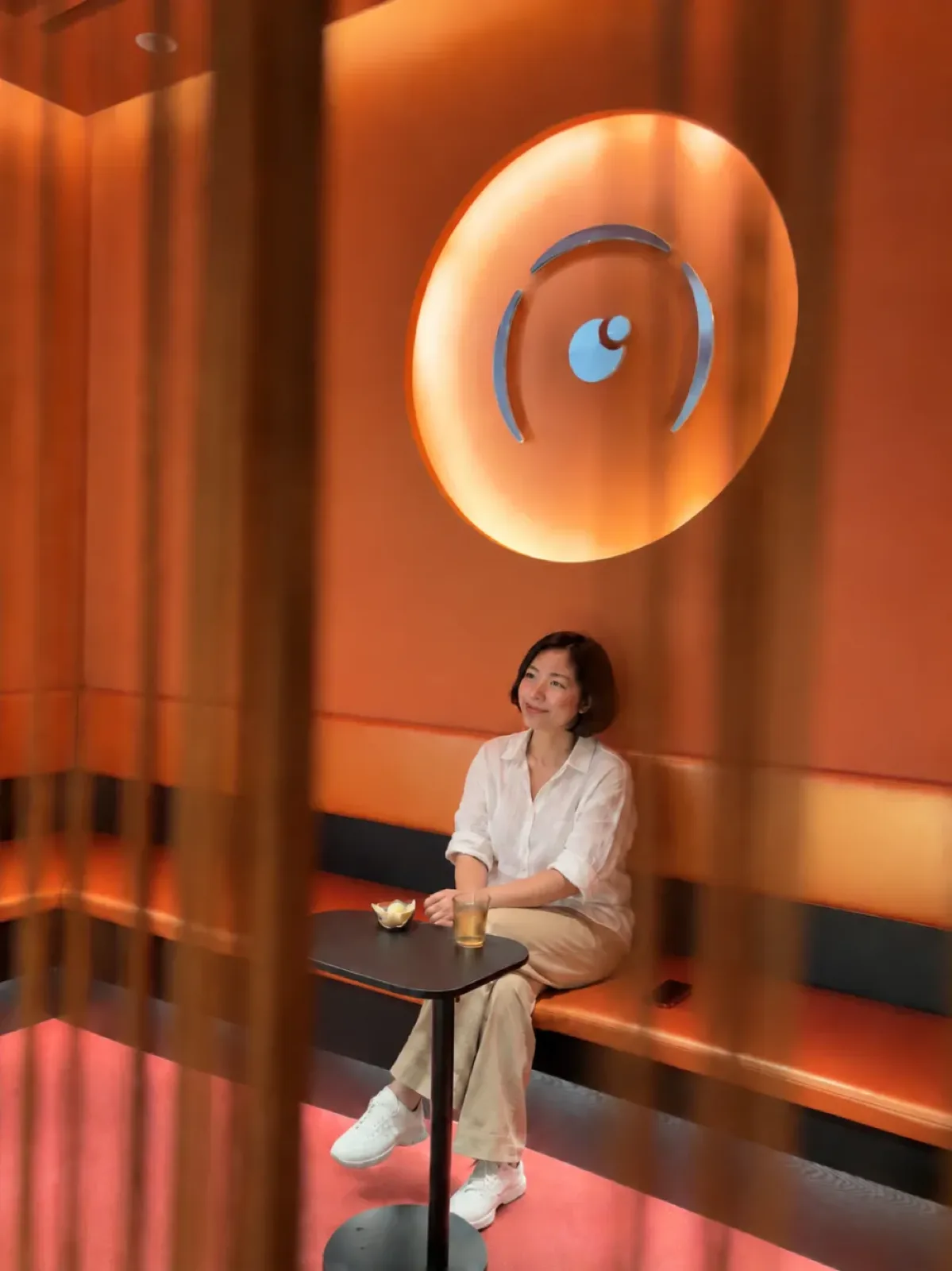 A woman in a white shirt sitting quietly on a long orange bench with a cup of tea, under a round glowing logo, seen through a curtain of thin cords