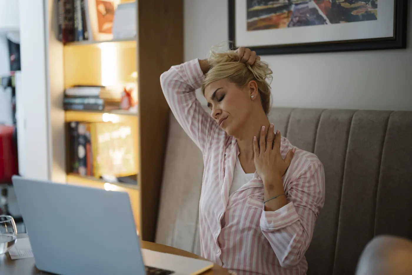 A woman at a laptop in a home office pausing to stretch her neck and shoulder